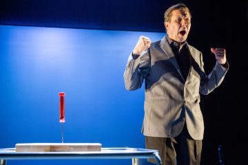 Harmaapukuinen mies seisoo nyrkit puristettuina ja näyttää huutavan tai laulavan sinisen taustan edessä. Punakahvainen veitsi pistetään pystyyn leikkuulautaan läheisellä pöydällä.