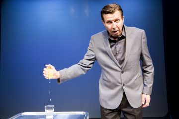 Harmaapukuinen mies puristaa puolikkaan sitruunan metallitarjottimella olevan vesilasin päälle, ja sitruunasta tippuu pisaroita. Hän katsoo eteenpäin yllättynyt ilme kasvoillaan ja seisoo tavallista sinistä taustaa vasten.