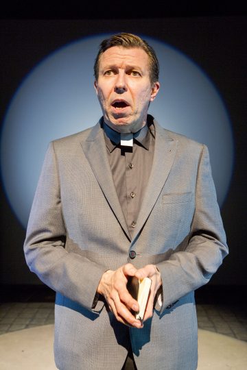 Harmaaseen pukuun pukeutunut papiksi pukeutunut mies seisoo valonheittimen alla, pitelee pientä kirjaa ja katsoo hieman ylöspäin ilmeikkäästi, suu auki.