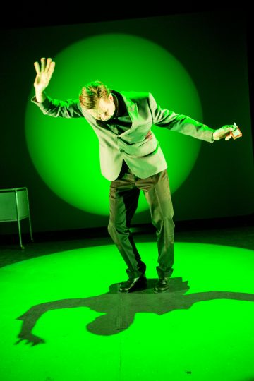 Pukuun pukeutunut henkilö kumartuu eteenpäin kirkkaan vihreässä näyttämövalaistuksessa pitäen toisessa kädessään pientä esinettä, jonka toinen käsi on nostettu ylös ja joka heittää dramaattisen varjon lattialle.