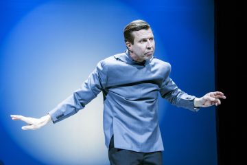 Siniseen pukutakkiin pukeutunut mies seisoo lavalla sinistä valonheitintä vasten, tekee dramaattisen ilmeen ja ojentaa kätensä ikään kuin korostaen jotakin asiaa tai näyttelemällä kohtausta.