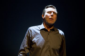 Tummaan napitettavaan paitaan pukeutunut mies seisoo dramaattisessa valaistuksessa mustaa taustaa vasten ja katsoo ylöspäin huolestuneena tai huolestuneena.