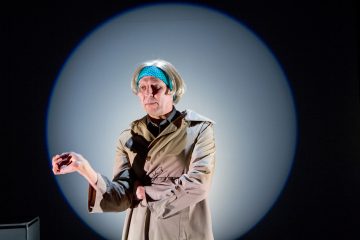 Henkilö, jolla on lyhyt harmaa peruukki, sininen otsapanta ja beige trenssitakki, seisoo valonheittimen alla tummalla näyttämöllä ja pitää yhtä kättä ojennettuna ja ristissä toista kättä.