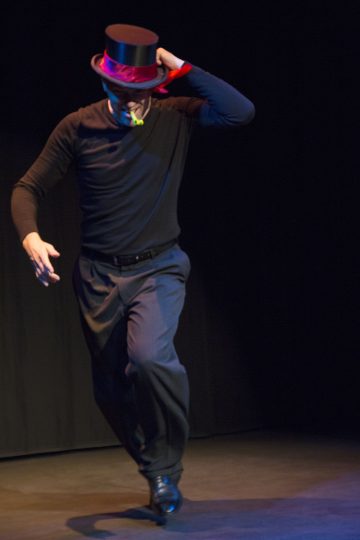 Mustaan silinterihattuun, mustaan paitaan ja mustiin housuihin pukeutunut henkilö tanssii energisesti lavalla toinen käsi kohotettuna ja toinen ojennettuna, ja hänet on kuvattu keskellä askelta tummaa taustaa vasten.