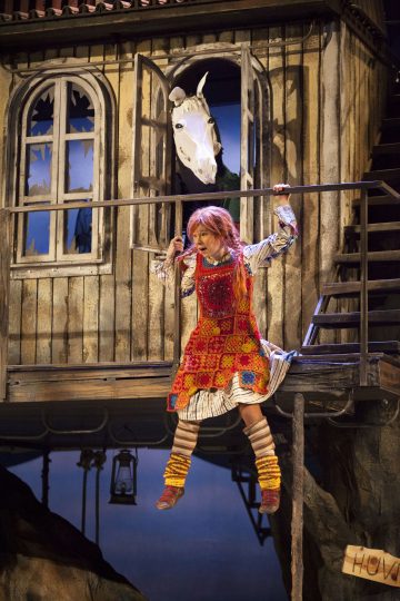 Pippi Pitkätossuksi pukeutunut henkilö roikkuu kaiteesta maalaismaisen puutalon edessä, kun valkoinen hevonen katsoo ulos yläkerran avoimesta ikkunasta hänen yläpuolellaan.