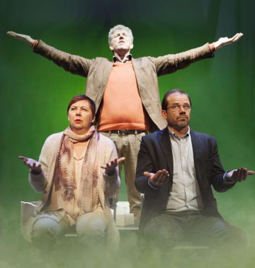 Lavalla kolme aikuista: mies seisoo takana kädet koholla ja silmät kiinni, kun taas nainen ja toinen mies istuvat edessä kädet avoinna, kämmenet ylöspäin, vihreää taustaa vasten.