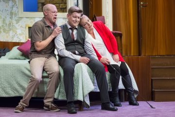 Kolme ihmistä istuu sängyn reunalla. Keskellä istuva, pukuun pukeutunut mies näyttää järkyttyneeltä, kun taas toinen mies ja nainen molemmin puolin lohduttavat häntä huolestunein ilmein viihtyisässä, matolla päällystetyssä huoneessa.