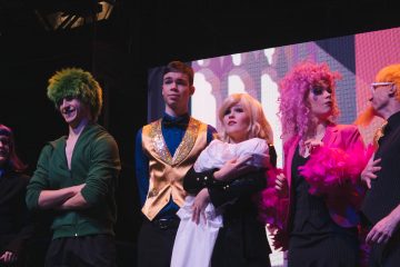 Lavalla seisoo viisi värikkäisiin teatteripukuihin pukeutunutta henkilöä. Yhdellä on vihreä peruukki, toisella kultainen liivi ja sininen rusetti, toisella vaaleat hiukset ja valkoinen liina kädessä, toisella vaaleanpunainen peruukki ja höyhenet ja yhdellä silmälasit.