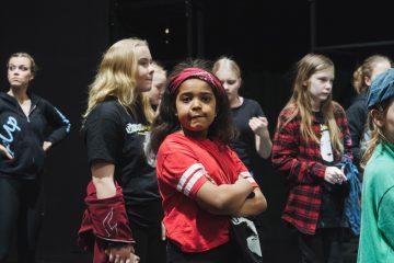 Punapaitainen ja otsapantainen tyttö seisoo itsevarmasti kädet ristissä muiden lasten joukossa, jotka ovat pukeutuneet rennosti ja näyttävät odottavan tai valmistautuvan johonkin tapahtumaan.