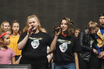 Kaksi nuorta naista laulaa lavalla mikrofoneihin, ja heillä on yllään samanlaiset mustat paidat, joissa on tyylitelty kasvokuva. Useita muita nuoria seisoo heidän takanaan, jotkut katsovat laulajia, toiset katsovat muualle.