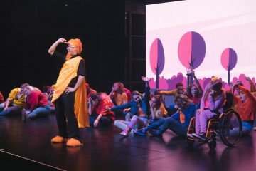 Lavalla on joukko nuoria esiintyjiä, joiden joukossa on myös pyörätuolissa istuva henkilö, värikkäissä puvuissa. Yksi oranssiin pukuun pukeutunut henkilö seisoo edessä, kun muut istuvat tai kurottautuvat ylöspäin, ja heidän takanaan on kirkas, tyylitelty puita esittävä tausta.