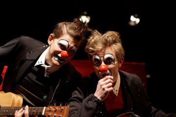 Kaksi pelleiksi pukeutunutta ihmistä, joilla on maalatut kasvot ja punaiset nenät, poseeraavat yhdessä, toinen pitelee kitaraa ja toinen mikrofonia, tummalla taustalla, jossa on lavavalot.