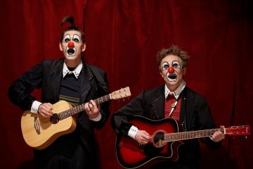 Kaksi pelleä, joilla on maalatut kasvot, punaiset nenät ja piikkitukka, soittaa akustisia kitaroita punaisen verhon edessä. Heillä on mustat takit ja ilmeikäs meikki, mikä luo leikkisän ja teatraalisen kohtauksen.