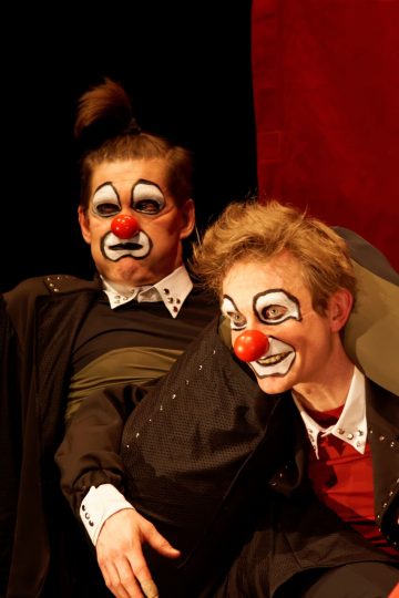 Kaksi klovneiksi pukeutunutta ihmistä, joilla on valkoiseksi maalatut kasvot, liioitellut ilmeet ja punaiset nenät, poseeraavat yhdessä teatteripuvuissa punaista verhoa vasten tummalla taustalla.