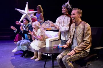 Lavalla istuu ja seisoo viisi värikkäisiin, omituisiin pukuihin pukeutunutta ihmistä, jotka katselevat tai hurraavat innoissaan. Yhdellä on kädessään suuri tähtisauva, toisella kruunu ja valtikka, ja he luovat eloisan, teatraalisen tunnelman kirkkaiden näyttämövalojen alla.