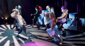 Kuusi näyttelijää, jotka ovat pukeutuneet hienostuneisiin, värikkäisiin pukuihin, esiintyvät energisesti näyttämöllä, jossa on dramaattinen valaistus, numeroiden ja hammaspyörien koristelemien lavasteiden edessä, mikä herättää omituisen, teatterillisen ilmapiirin.