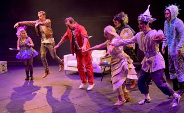 Ryhmä värikkäisiin pukuihin pukeutuneita näyttelijöitä esiintyy energisesti lavalla, jotkut kädet ojennettuina tai kesken hypyn, valkoisen sohvan ja rekvisiitan edessä dramaattisessa valaistuksessa.