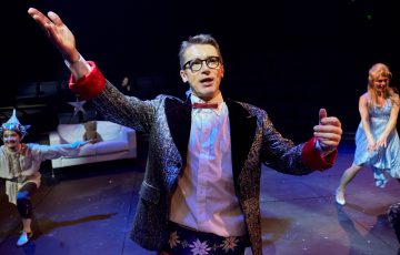 Mies, jolla on kimalteleva bleiseri ja silmälasit, elehtii dramaattisesti lavalla, ja taustalla tanssii kaksi juhlapukuista esiintyjää teatterivalaistuksessa.