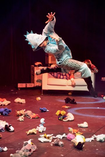 Esiintyjä hyppää dramaattisesti näyttämölle, jota ympäröivät hajallaan olevat pehmolelut, ja taustalla on valkoinen sohva ja värikkäitä rekvisiittaa näyttämövalaistuksen alla.