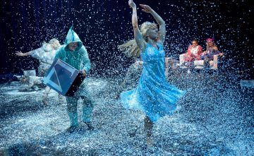 Vaaleansiniseen mekkoon pukeutunut nainen tanssii iloisesti keinolumen sattuessa. Lähistöllä siniseen asuun pukeutunut henkilö pitelee laatikkoa, ja taustalla istuu tai leikkii muita, jotka kaikki ovat putoavan lumen ympäröimiä.