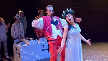 Vaaleansiniseen mekkoon ja kruunuun pukeutunut nainen laulaa dramaattisesti lavalla kädet ojennettuina, kun punatakkinen mies seisoo hänen takanaan ja tekee ilmeikkään eleen. Taustalla näkyy muita esiintyjiä ja valkoinen laatikko, jossa on merkintä 24.