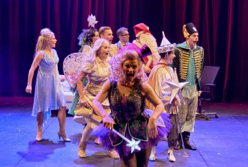 Ryhmä värikkäisiin, omituisiin pukuihin pukeutuneita esiintyjiä seisoo lavalla, jossa on tummanpunaiset verhot; edessä olevalla naisella on violetti tutu ja kädessään taikasauva, kun taas muilla on keijujen, sotilaiden ja satuhahmojen asuja.