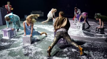 Ryhmä värikkäisiin pukuihin pukeutuneita esiintyjiä liikkuu ja poseeraa tarmokkaasti valkoisella konfetilla päällystetyllä näyttämöllä ja on vuorovaikutuksessa numeroitujen harmaiden ja valkoisten palikoiden kanssa dramaattisessa valaistuksessa.