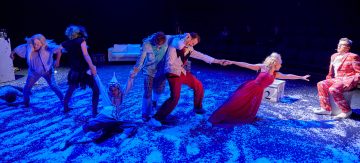 Ryhmä värikkäisiin pukuihin pukeutuneita esiintyjiä poseeraa dramaattisesti lavalla, jonka lattia on peitetty valkoisella konfetilla. Kaksi punaisiin pukeutunutta ihmistä kurottautuu toisiaan kohti, kun taas toiset vetävät heidät erilleen sinisen näyttämövalaistuksen alla.