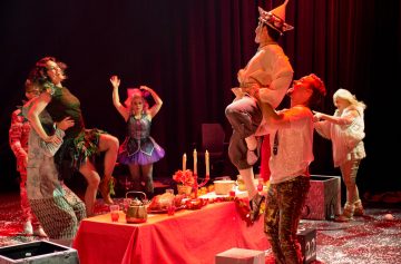 Vauhdikkaassa näyttämöteoksessa pukuun pukeutuneet näyttelijät tanssivat ja juhlivat juhlallisesti koristellun pöydän ympärillä, ja yksi näyttelijä nostaa toista näyttelijää ilmaan värikkään näyttämövalaistuksen alla.