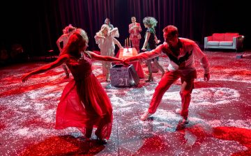 Joukko juhlapukuisia ihmisiä tanssii tarmokkaasti valkoisilla konfeteilla päällystetyllä lavalla, jota valaisee dramaattinen punainen valaistus. Taustalla on pöytä kynttilöineen ja sohva.