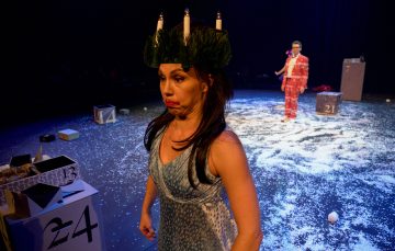 Siniseen mekkoon pukeutunut nainen, jolla on kynttiläkruunu, tekee lavalla hassun naaman. Taustalla punapukuinen mies seisoo numeroitujen laatikoiden ja keinolumen ympäröimänä näyttämövalojen alla.