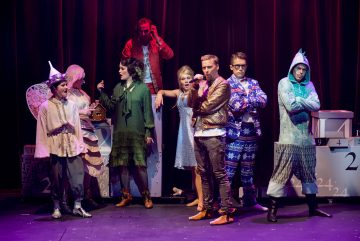 Kahdeksan värikkäisiin, omituisiin pukuihin pukeutuneen ihmisen ryhmä seisoo lavalla punaisen verhon edessä ja poseeraa dramaattisesti suurten numeroitujen korttelien keskellä. Jotkut näyttävät yllättyneiltä, toiset itsevarmoilta tai mietteliäiltä.