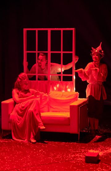 Kolme näyttelijää esiintyy lavalla punaisen valaistuksen alla. Yksi istuu valkoisella sohvalla kaapu yllään, toinen nojaa sohvan takana olevasta ikkunanpuitteesta, ja kolmas seisoo sivussa teräväkärkinen hattu ja puku yllään.