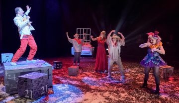 Viisi värikkäisiin teatteripukuihin pukeutunutta ihmistä esiintyy energisesti tekolumen peittämällä lavalla, ja yksi henkilö laulaa korotetulla korokkeella, kun muut poseeraavat dramaattisesti. Suuria numeroituja laatikoita on ripoteltu ympäri lavaa.