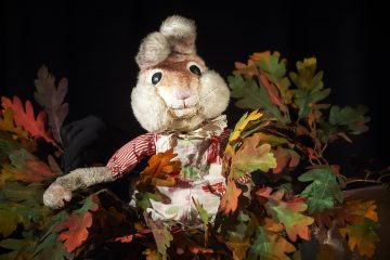 Kulunut, pehmoinen jänisnukke, jolla on punavalkoinen paita ja esiliina, on värikkäiden keinotekoisten syyslehtien ympäröimä tummalla taustalla.