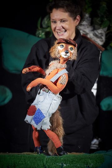 Hymyilevä mustapukuinen nukketeatterinjohtaja ohjaa oranssia ketunukkea, joka on pukeutunut laastaroituihin haalareihin, suojalaseihin ja punaisiin kenkiin.