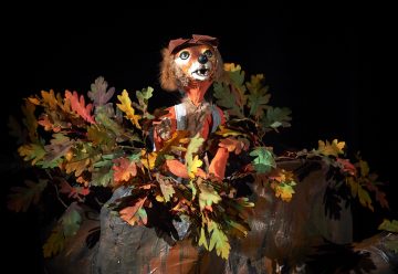 Kettu-nukke, jolla on punainen lippis ja haalarit, seisoo värikkäiden syyslehtien keskellä tummalla taustalla.