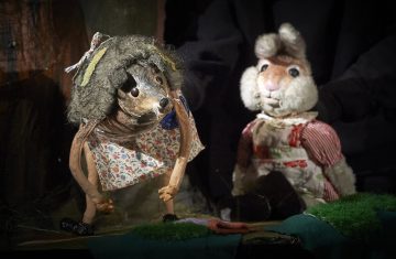 Lavalla on kaksi eläinnukkea: siili, jolla on kukkamekko ja pörröinen pää, ja kani, jolla on punaraitainen mekko ja valkoinen esiliina. Mustiin pukeutunut henkilö operoi nukkeja hämärästi valaistussa kohtauksessa.