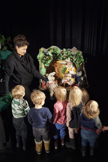 Nainen esittää nukketeatteriesitystä eläinnukeilla metsäaiheisessa lavastuksessa, kun ryhmä pieniä lapsia seisoo edessä ja katsoo tarkkaavaisesti.