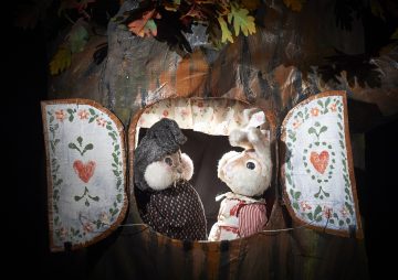 Kaksi pehmonukkea, mustatukkainen hahmo mekossa ja valkoinen kani raidallisessa asussa, ovat vastakkain ikkunassa, jota kehystävät maalatut kukkaverhot ja jota ympäröivät lehdet.