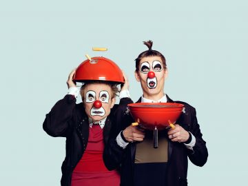 Kaksi pelleiksi pukeutunutta ihmistä, joilla on valkoinen kasvomaali ja punainen nenä; toinen pitää päässään punaista grilliritilän kantta ja toinen grilliritilän pohjaa leuan alla, molemmilla yllättyneet ilmeet, vaaleaa taustaa vasten.