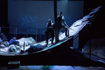 Näyttelijät ovat hämärästi valaistussa lavastuksessa, joka muistuttaa kallistunutta, sään vaikutuksesta kulunutta laivaa; kaksi seisoo kiväärien kanssa, kun taas toiset makaavat tai istuvat kaltevalla kannella, mikä luo dramaattisen ja jännittyneen ilmapiirin.