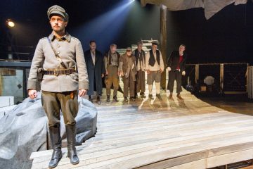 Sotilaspukuinen mies seisoo puisen näyttämön etuosassa, kun taas hänen takanaan seisoo rivissä kuuden vaihteleviin vaatteisiin pukeutuneen miehen ryhmä dramaattisessa valaistuksessa teatteriympäristössä.