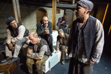 Kuusi kuluneisiin, vanhanaikaisiin vaatteisiin pukeutunutta miestä istuu ja seisoo näyttämöllä, joka muistuttaa maalaismaista, hämärästi valaistua huonetta. He näyttävät keskustelevan syvällisesti, ja heidän ilmeensä ovat vakavia ja mietteliäitä.