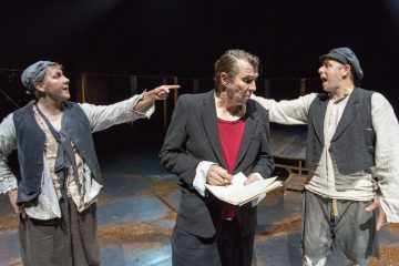 Lavalla esiintyy kolme rähjäisiin vaatteisiin pukeutunutta näyttelijää, joista kaksi seisoo molemmin puolin osoittaen ja huutaen keskellä olevaa miestä, joka katsoo alaspäin pitäen kädessään papereita ja kynää. Tausta on hämärästi valaistu.