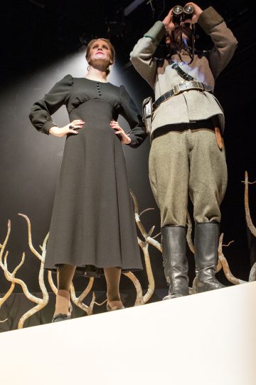 Pitkään mustaan mekkoon pukeutunut nainen seisoo itsevarmasti harmaaseen sotilaspukuun ja saappaisiin pukeutuneen henkilön vieressä, joka katsoo kiikareilla. Taustalla on taiteellisia oksia, ja kohtaus on dramaattisesti valaistu.