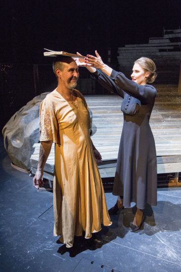 Mustapukuinen nainen asettaa kirjan hymyilevän, persikanväriseen mekkoon pukeutuneen miehen päähän hämärästi valaistulla näyttämöllä, jonka taustalla on pyöreä puinen alusta ja suuri kivi.