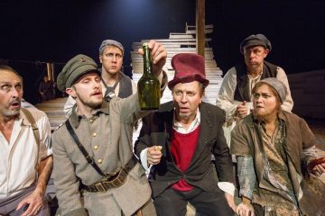 Kuusi rähjäisiin aikakauden pukuihin pukeutunutta näyttelijää istuu tiiviisti yhdessä näyttämöllä ja katselee keskittyneesti vihreää lasipulloa, jota yksi heistä pitää ylhäällä, osoittaen yllätyksen ja uteliaisuuden yhdistelmää puista kulissia vasten.