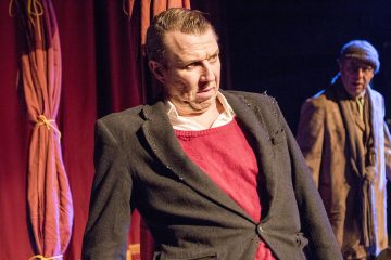 Mustaan pukuun ja punaiseen villapaitaan pukeutunut mies nojaa lavalla kömpelösti ja yllättyneenä, kun taas toinen lippalakkipäinen mies seisoo taustalla, lähellä punaisia verhoja.