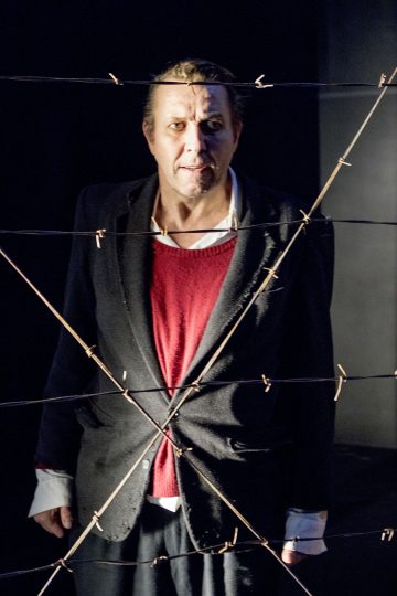 Tummaan bleiseriin ja punaiseen villapaitaan pukeutunut mies seisoo piikkilanka-aidan takana ja katsoo eteenpäin. Valaistus on dramaattinen, ja varjot korostavat miehen vakavaa ilmettä.